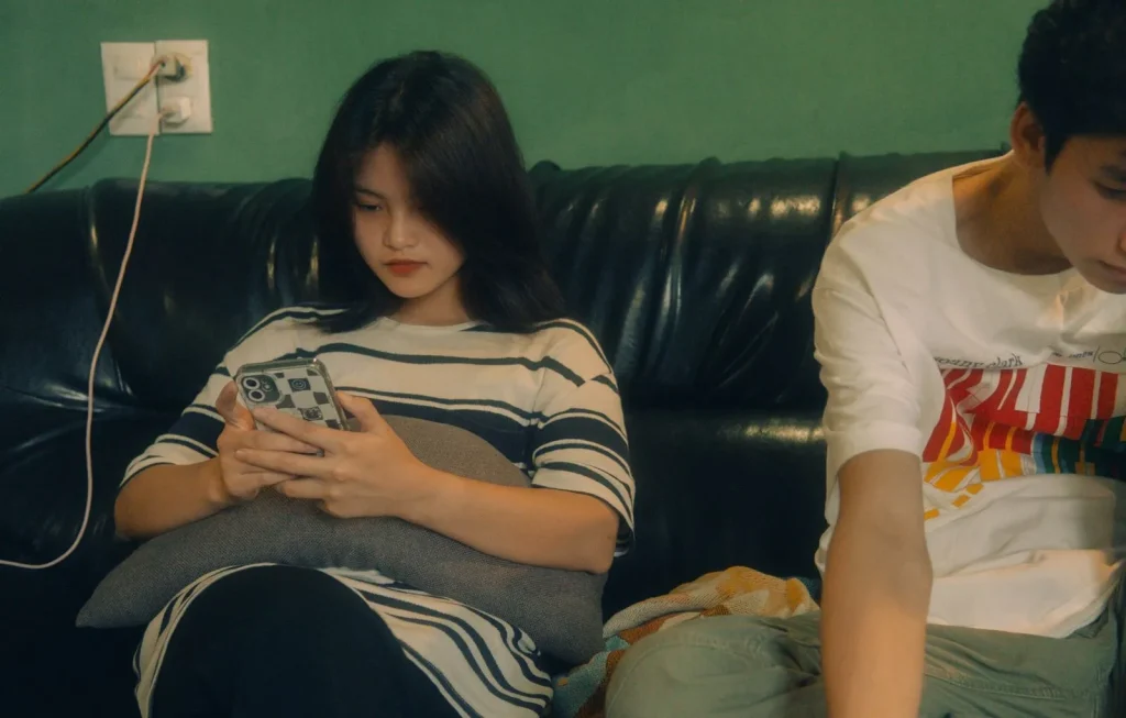 Two teenagers sit together on a sofa, each focused on a smartphone.